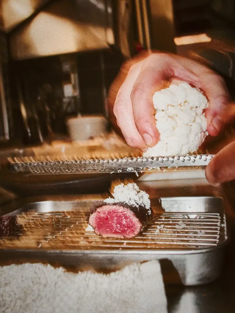 Main râpant du chou-fleur sur une pièce de viande rosée – détail gastronomique par Jonathan Thevenet à Lyon