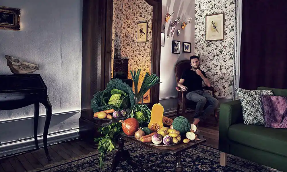 Portrait du photographe culinaire Jonathan Thevenet assis dans un intérieur vintage, avec une nature morte de légumes de saison en premier plan.