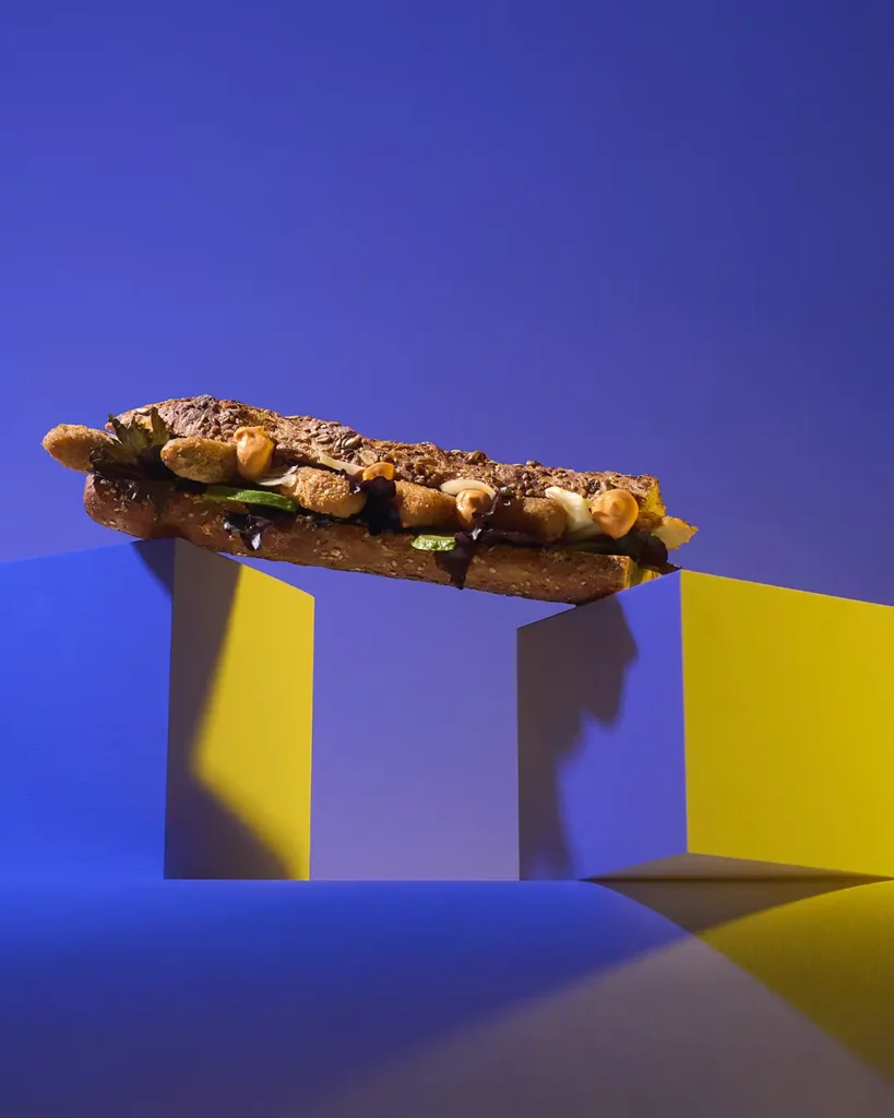 Sandwich gastronomique aux nuggets et pousses fraîches, posé en lévitation sur des volumes géométriques bleu et jaune — photographie culinaire premium
