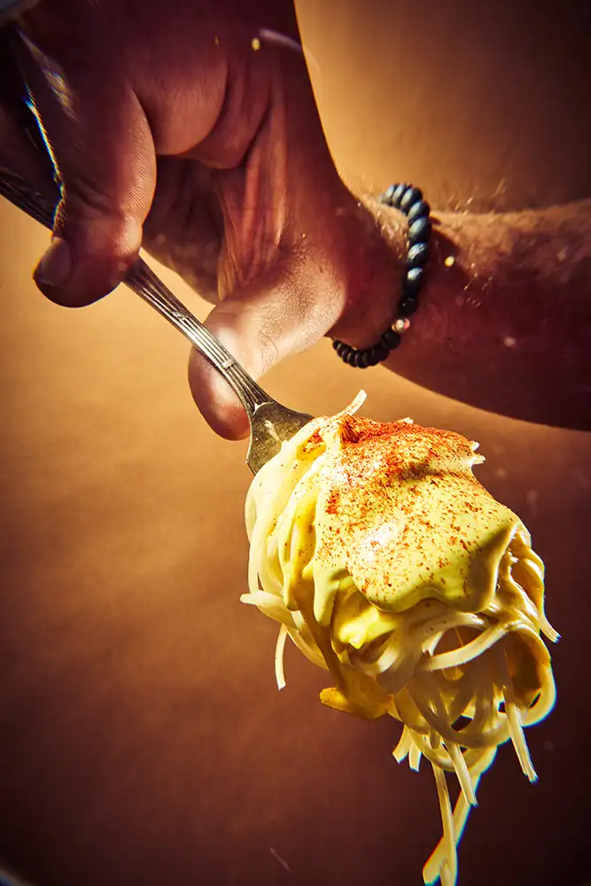Main tendant une fourchette de pâtes crémeuses saupoudrées de paprika sur fond ocre chaud — série culinaire « Mange ! », photographe culinaire à Lyon.