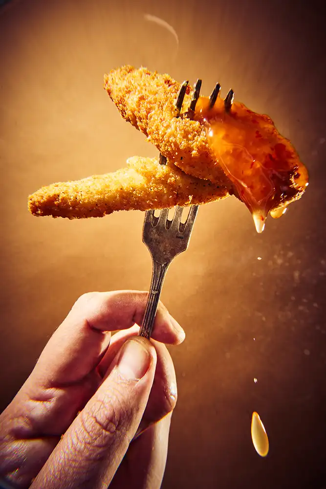 Main tendant une fourchette d'aiguillettes de poulet panées nappées de sauce aigre-douce qui coule, fond ocre — série culinaire « Mange ! », photographe culinaire à Lyon.
