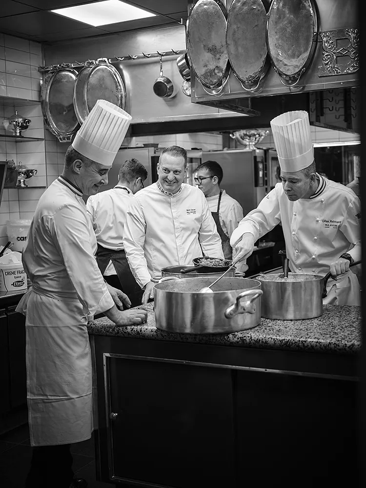 Reportage en noir et blanc dans les cuisines du restaurant Paul Bocuse, brigade au travail autour des sautoirs, photographe culinaire Lyon
