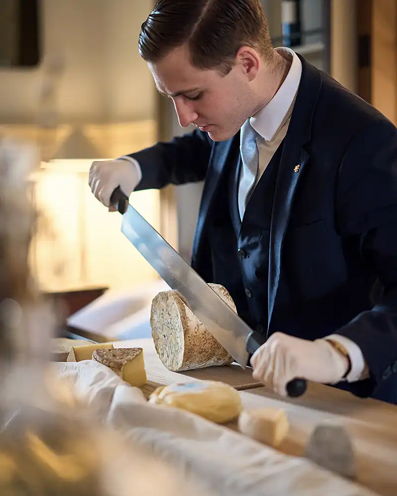 Chef de rang en gants blancs découpant un fromage affiné au guéridon, lumière chaude latérale, restaurant Saisons, photographe culinaire Lyon