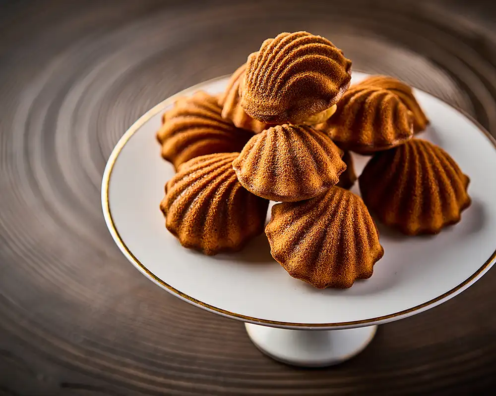 Madeleines dorées dressées sur un présentoir, fond de bois, lumière chaude, restaurant Saisons, photographe culinaire Lyon