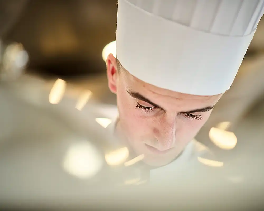 Portrait serré d'un jeune cuisinier en toque, penché sur son travail, reflets de cloche, restaurant Saisons, photographe culinaire Lyon