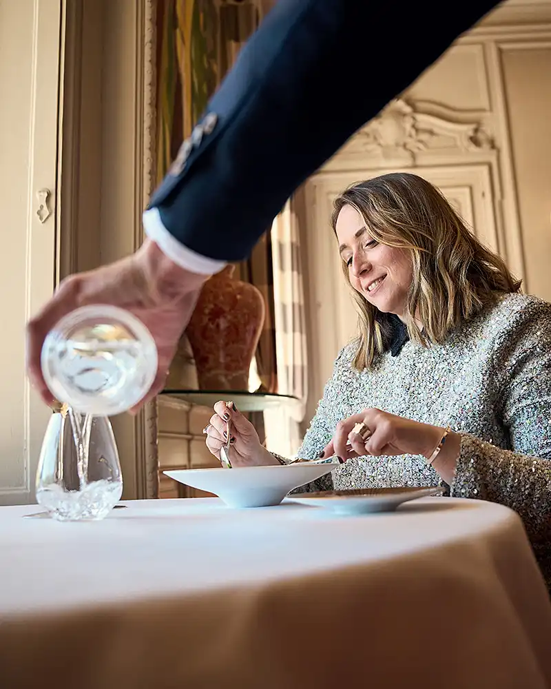 Maître d'hôtel versant de l'eau à une cliente souriante en salle du restaurant Saisons, lumière naturelle, photographe culinaire Lyon
